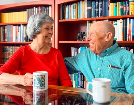 Two smiling seniors sit at a glass table, talking over coffee mugs with colorful bookshelves behind them in a library setting.