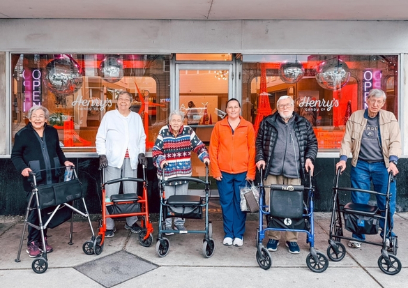 Seven seniors pose with their rolling walkers outside a storefront with red decor and open signs, in front of a cafe called Henry's.