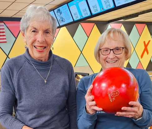 Two smiling elderly women at a bowling alley; one holds a bright red bowling ball, ready to bowl.