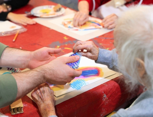 Close-up of hands guiding an elderly person painting a blue patterned cup at a colorful craft table during an arts-and-crafts session.