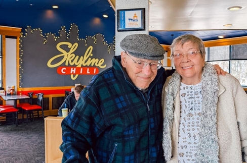 Two smiling elderly people posing together inside Skyline Chili; man wears a flat cap and plaid jacket, woman wears a light cardigan with a scarf, with the restaurant's chili sign in the background.