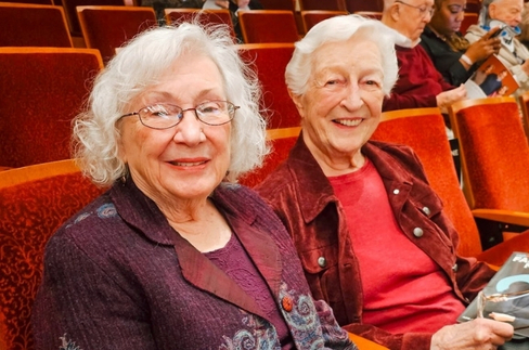 Two smiling elderly women seated in red theater seats, wearing jackets.