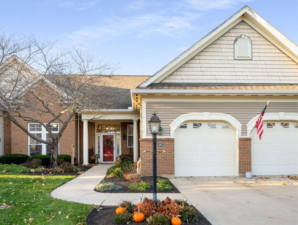 Suburban brick house with a white garage, front porch, and pumpkins arranged in the yard for autumn decorating, with a leafless tree nearby and an American flag by the garage door.