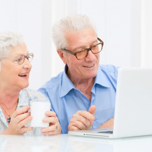 Happy senior couple looking at a laptop together; the man points at the screen while the woman holds a mug at a bright white table.