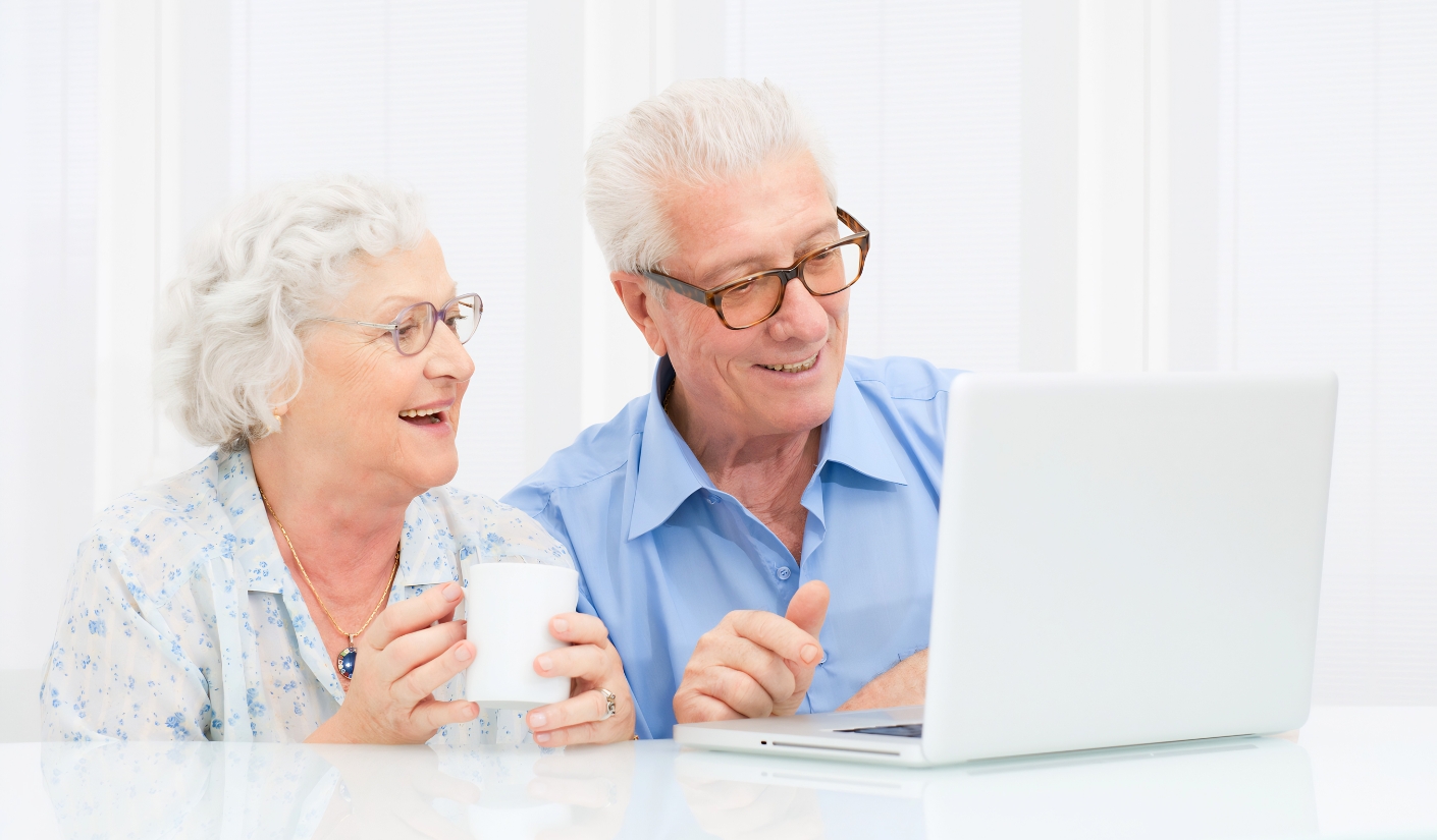 Happy senior couple looking at a laptop together; the man points at the screen while the woman holds a mug at a bright white table.