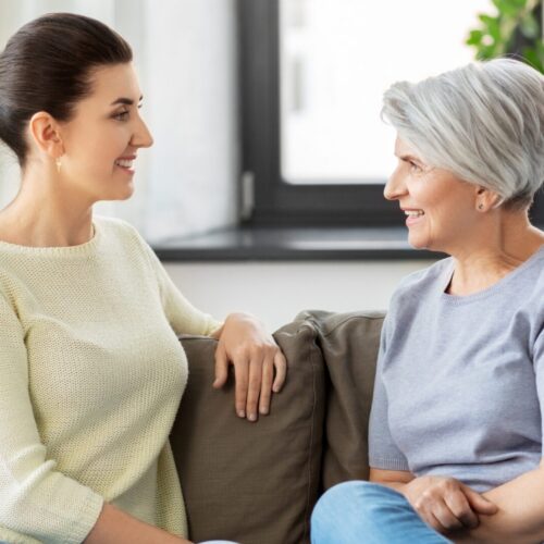Two women sit on a sofa facing each other, smiling in a bright living room setting.3e0 They appear to be enjoying a friendly conversation on a couch with patterned pillows nearby.4843a8